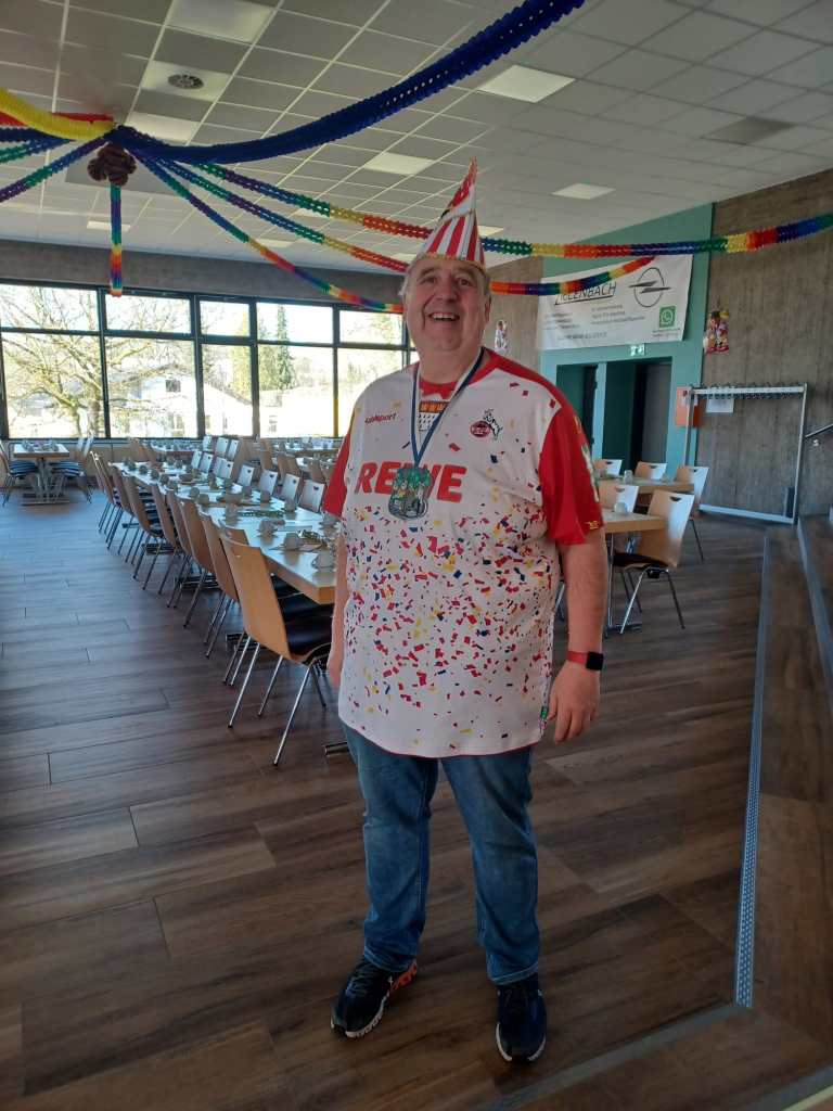 Ein Mann steht im Raum und hinter ihm sind Tische und Stühle. Alles ist bunt geschmückt und der Mann trägt einen bunten Hut, sowie in rotes T-Shirt.