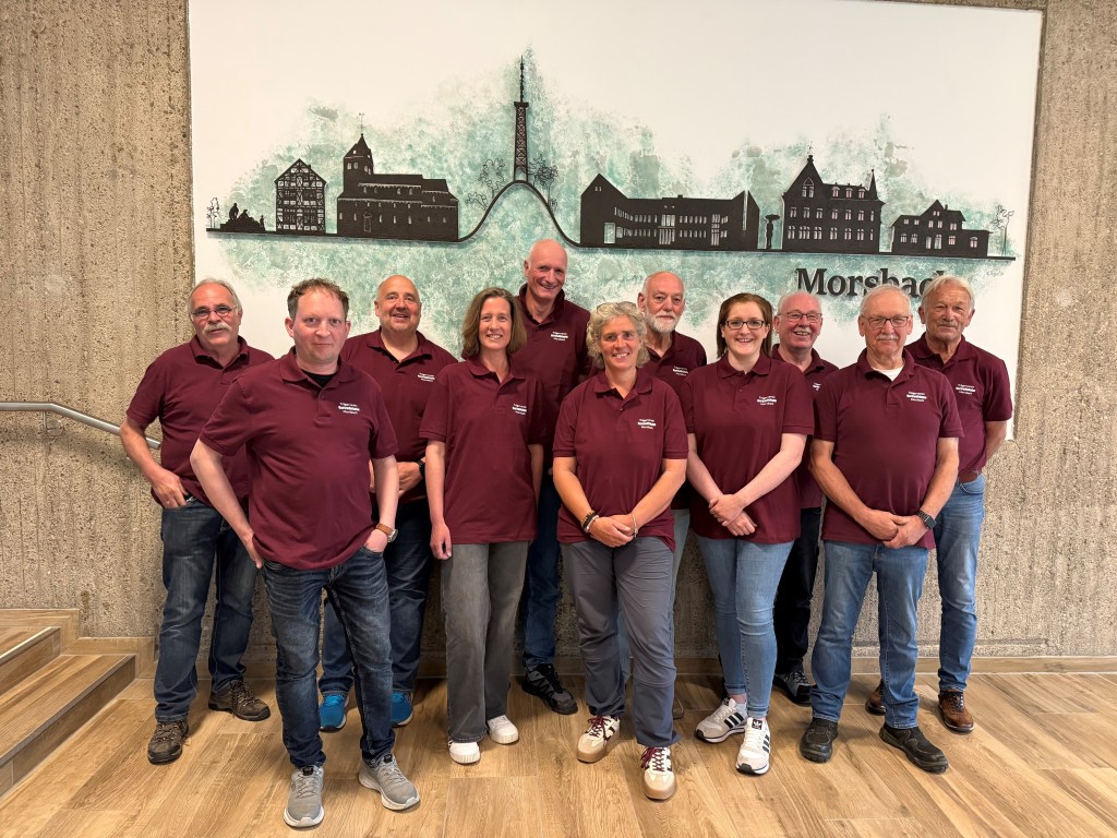 Eine Gruppe von elf Personen steht nebeneinander in einem Innenraum mit Holzfußboden und strukturierter Wand. Alle tragen einheitliche bordeauxrote Poloshirts und Jeans oder Freizeithosen. Hinter ihnen ist ein großes Wandbild mit stilisierten Gebäuden und einem Turm sowie dem Schriftzug „Morsbach“ zu sehen. Die Personen lächeln in die Kamera und stehen locker in zwei leicht versetzten Reihen.